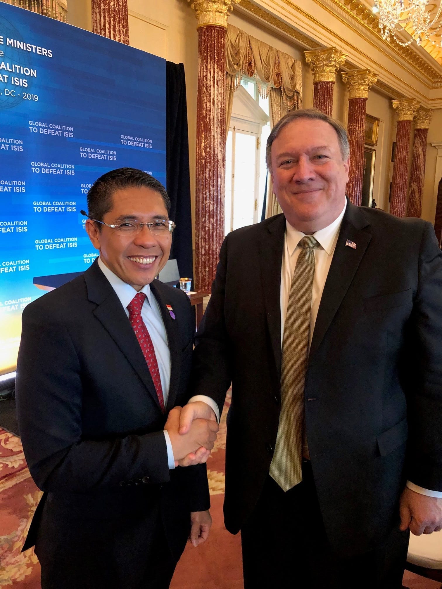 Two men in suits shaking hands in front of a "Global Coalition to Defeat ISIS" banner. One is Mike Pompeo.
